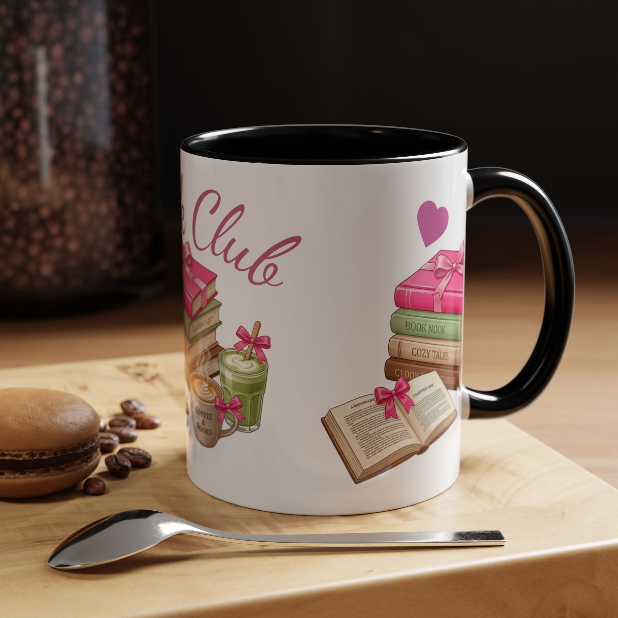 A white ceramic coffee mug with a black interior and handle, decorated with book-themed illustrations and the word 'Club' in pink script. The mug sits on a wooden surface with a spoon nearby, with a blurred background showing coffee beans and a macaron.