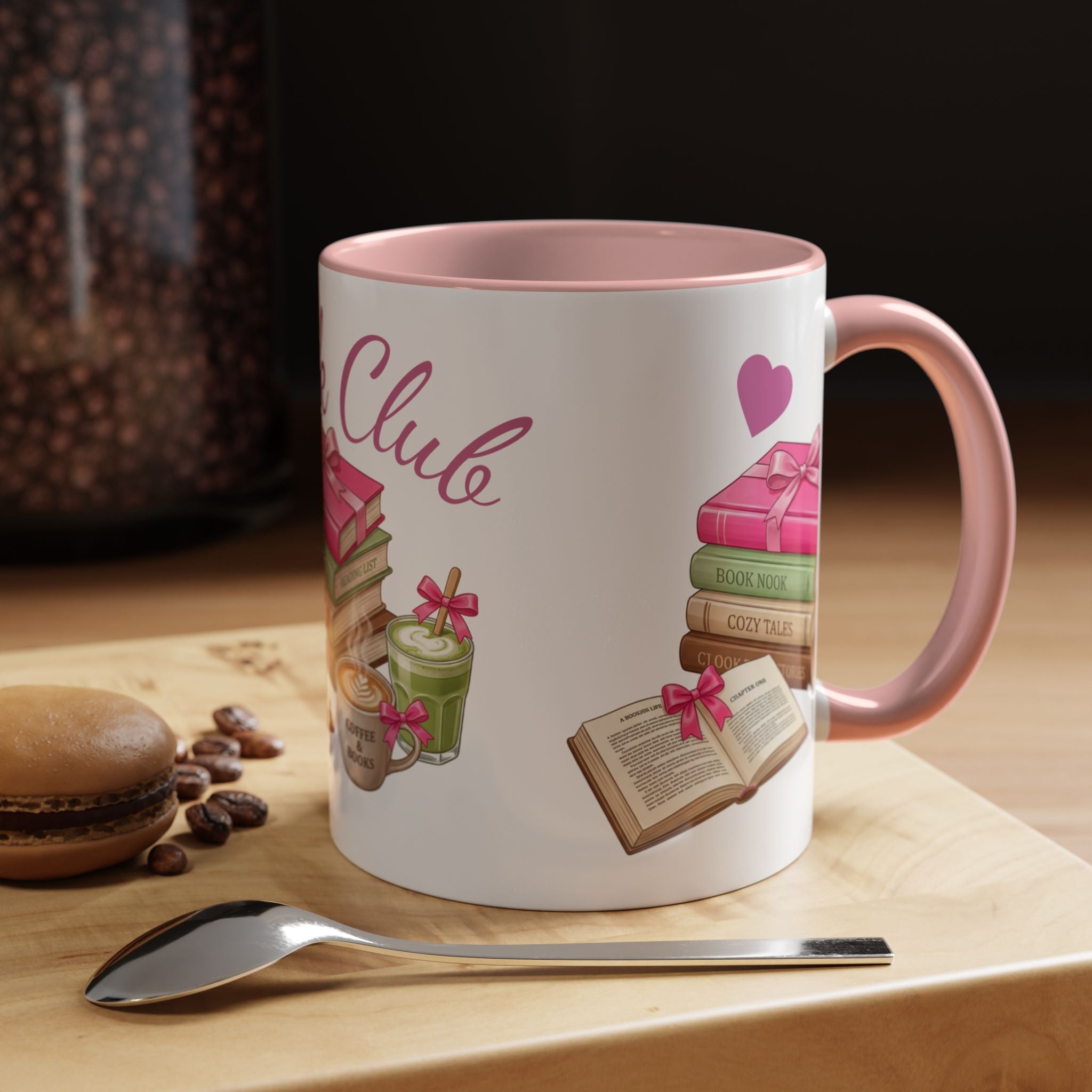 A white ceramic coffee mug with a pink interior and handle, decorated with illustrations of books, a heart, and the words 'Book Club'. The mug is set on a wooden surface next to a spoon, with a blurred background showing coffee beans and a macaron.