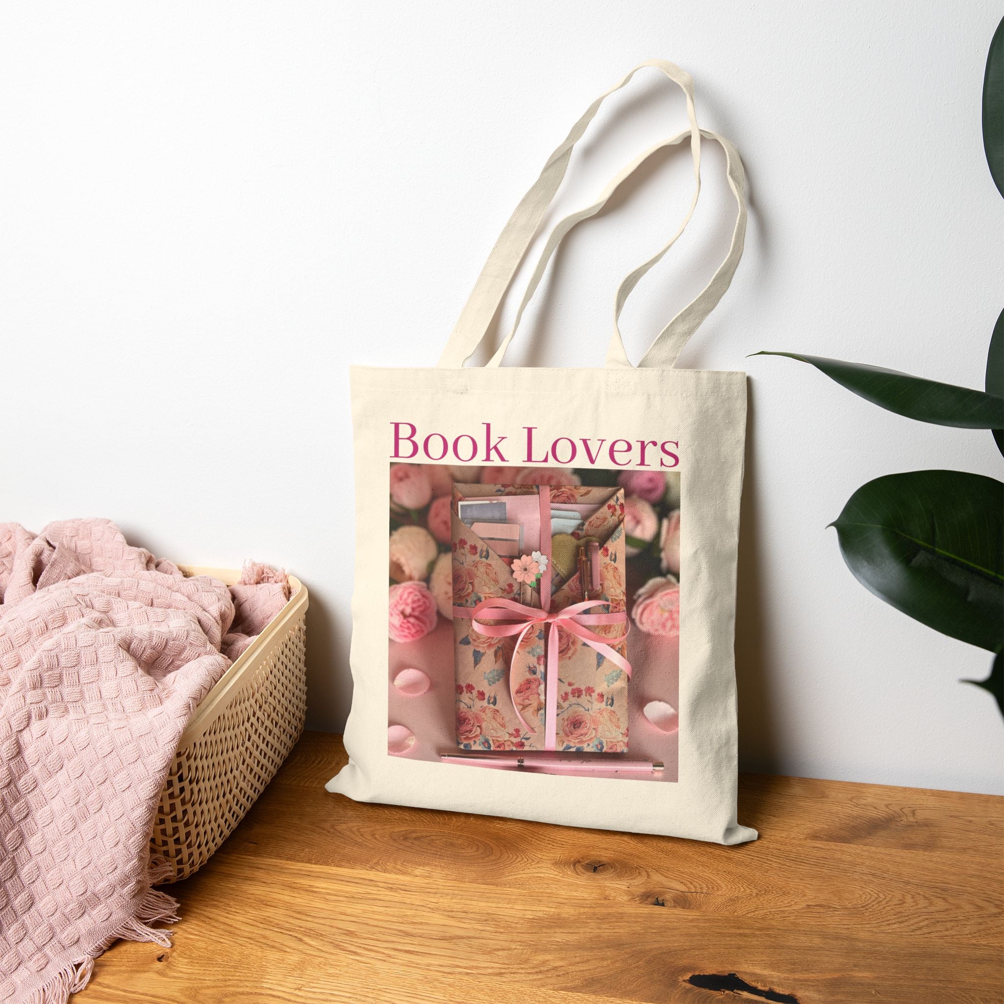 A cream canvas tote bag with 'Book Lovers' printed in pink text, featuring a central graphic of a floral book with a pink ribbon. The bag is styled on a wooden table next to a pink blanket in a basket and a green plant against a white wall.