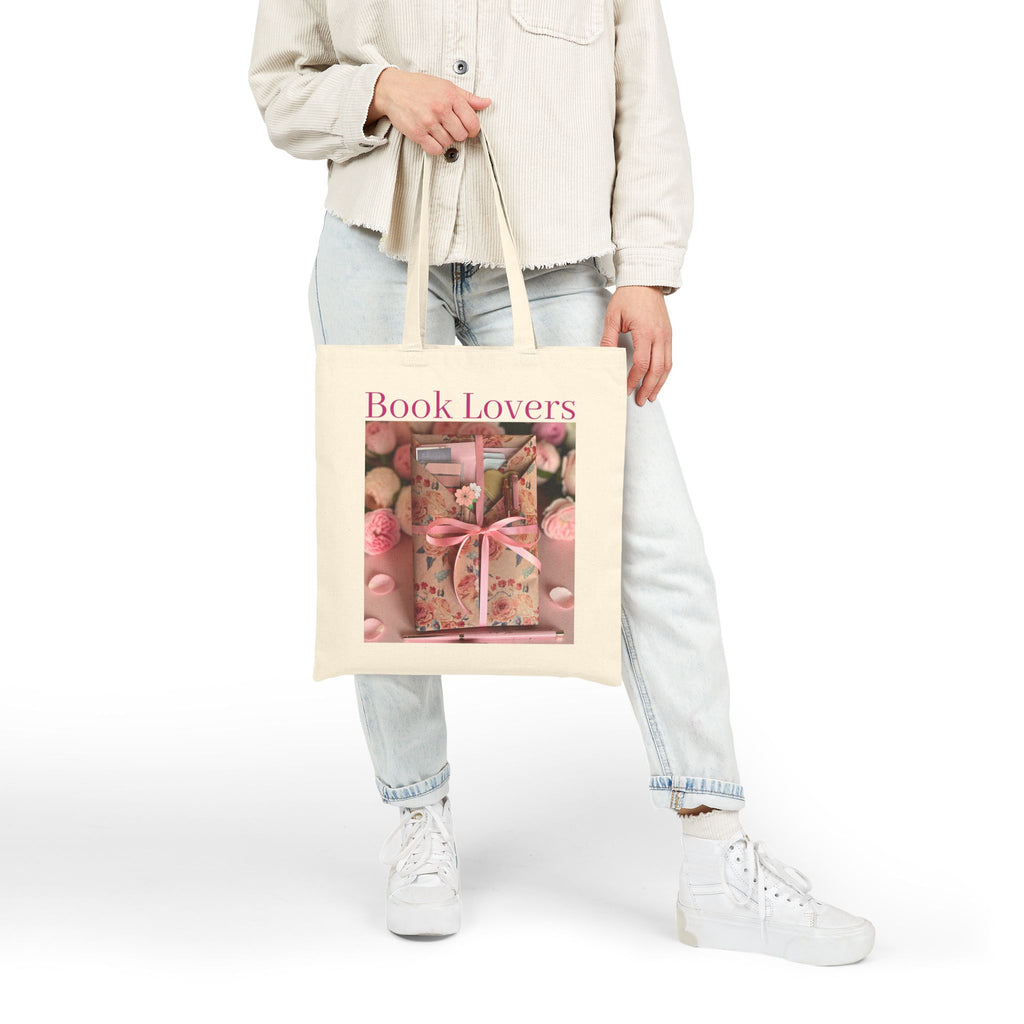 A person wearing a light beige jacket, light wash jeans, and white sneakers holds a cream canvas tote bag. The bag features a graphic print of a pink book box with a pink bow and the words 'Book Lovers' in pink text.