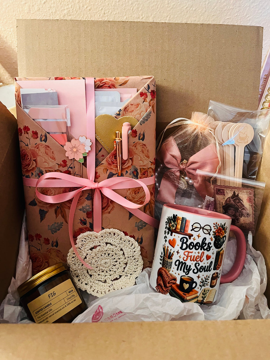 A curated gift box filled with stationery and cozy accessories, including a floral wrapped book tied with a pink ribbon, a pink mug that reads 'Books Fuel My Soul,' a small jar candle, a lace doily, and packaged wooden accessories.
