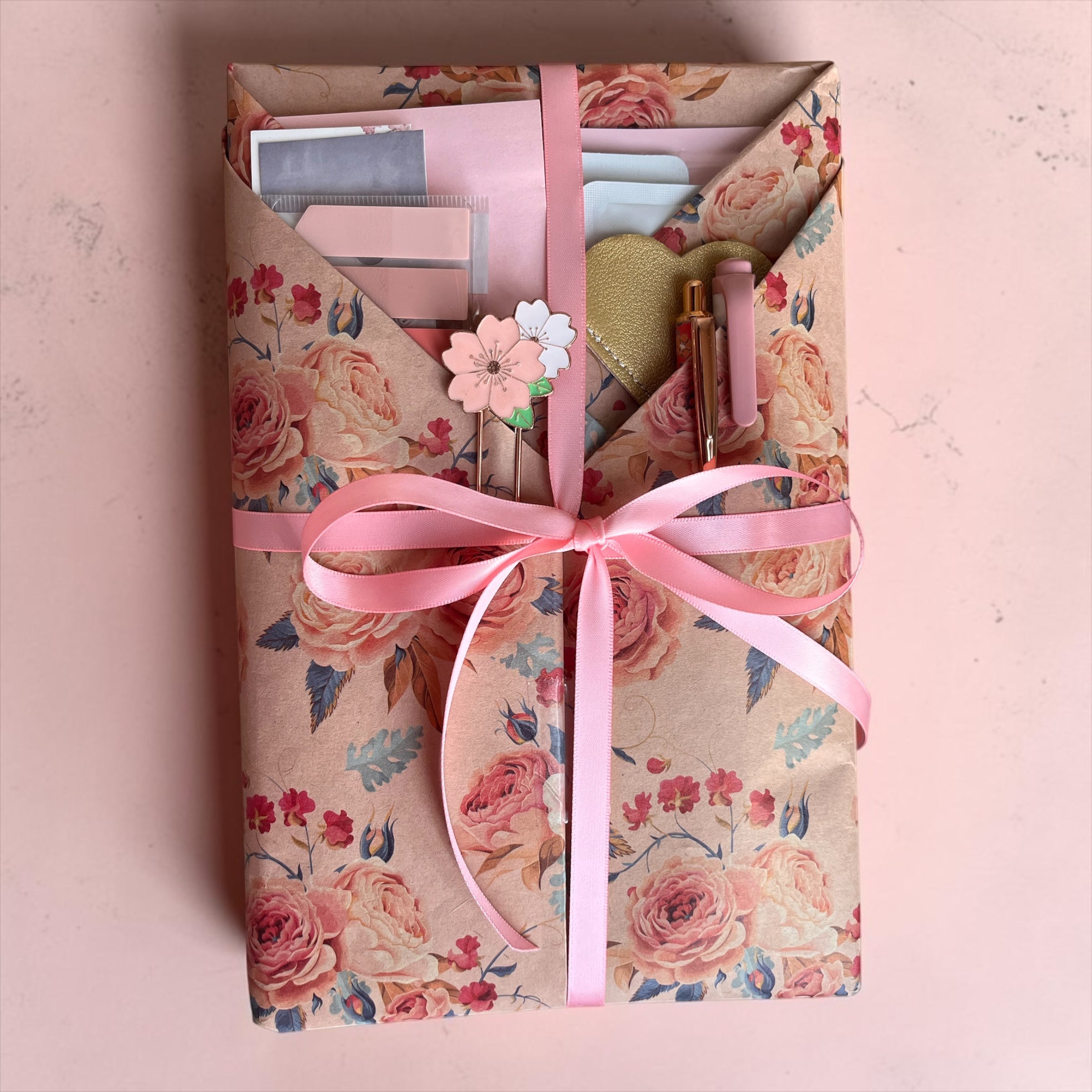 A gift box wrapped in pink floral paper with a pink ribbon bow, shown from above on a pale pink background. The box contains stationery items including page tabs, a pen, and a small flower-shaped pin. Also a lip, face mask and mud mask is included. 