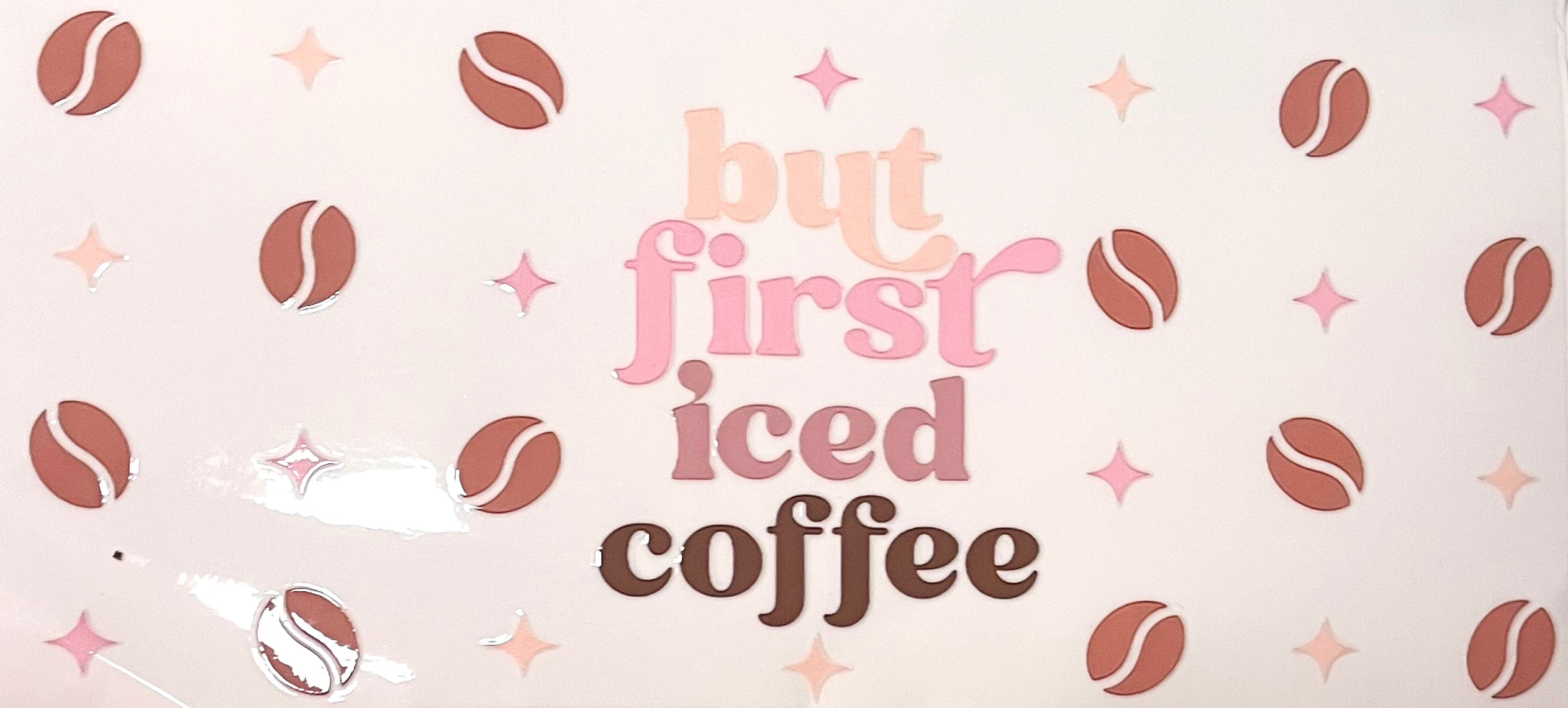 A Libby glass cup pattern with the main focus of the words “but first ice coffee,” at the center of the design. The words contain the color tones of light brown, pink, mauve and brown. Scattered around the words are pink and light brown starts mixed with various brown cartoonish coffee beans. 