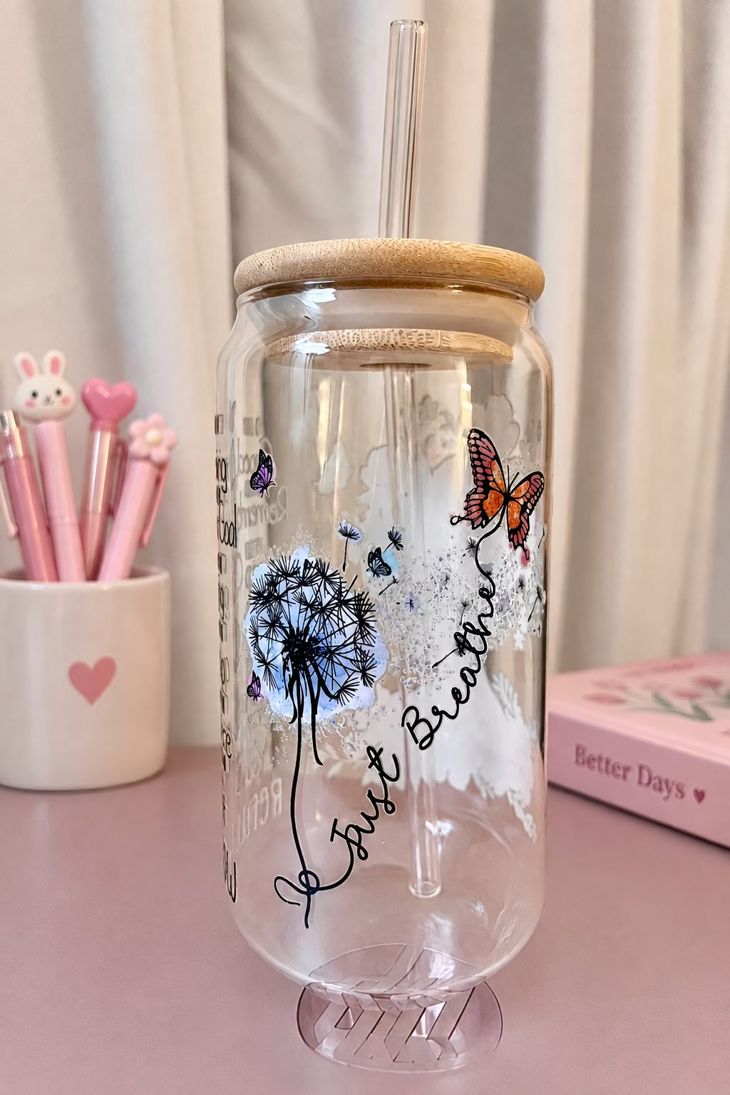 A clear glass can-shaped tumbler with a bamboo lid and clear straw, decorated with a blue dandelion, an orange butterfly, and the words 'Just Breathe' in black script, set against a soft beige curtain background.