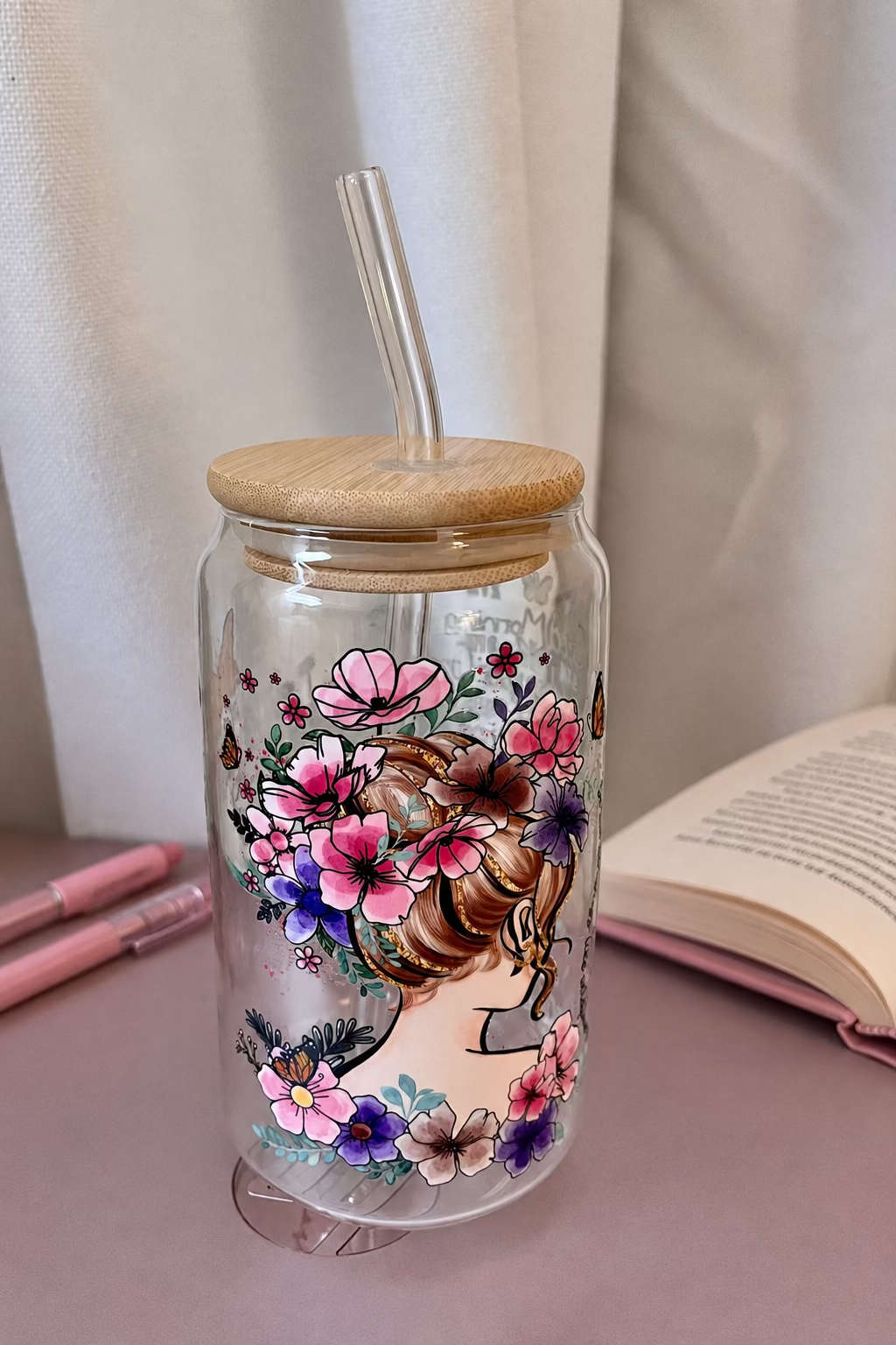 A clear glass tumbler with a bamboo lid and clear straw, decorated with a colorful illustration of a woman's profile surrounded by pink, purple, and blue flowers.