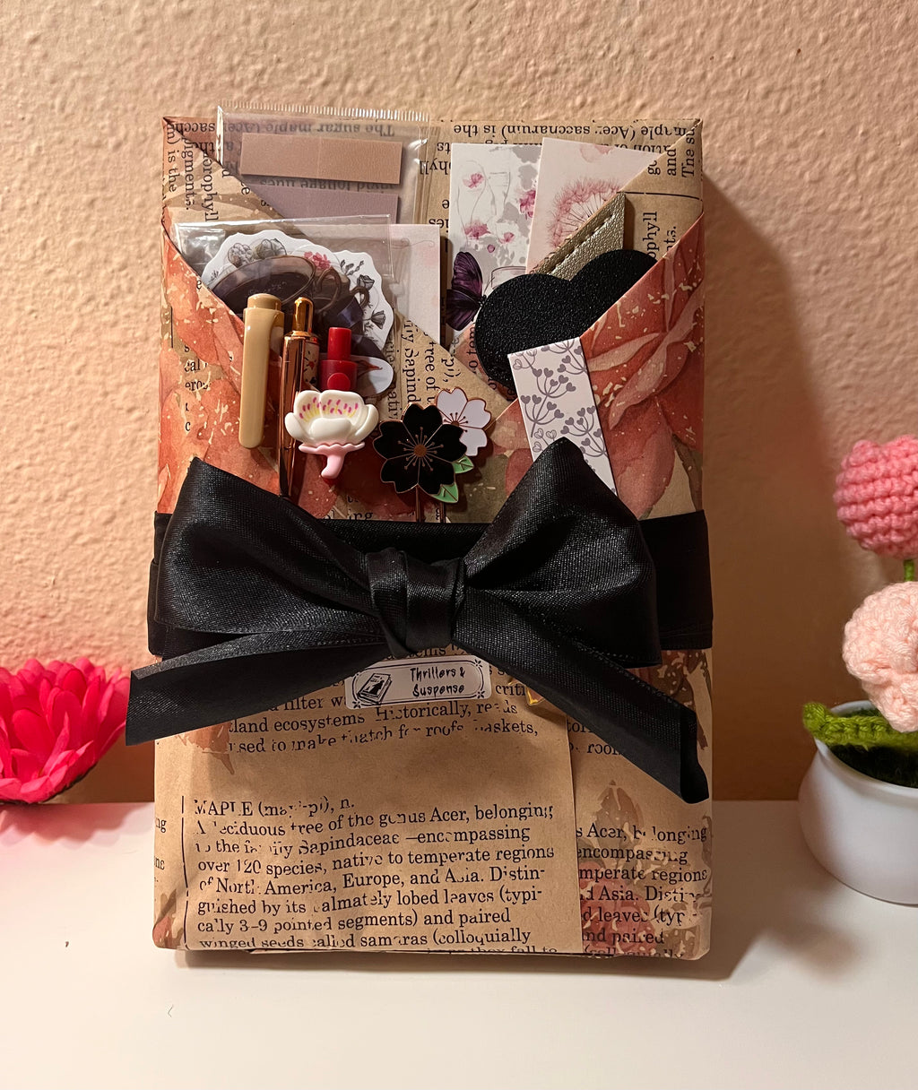 A wrapped Blind date with a book featuring vintage-style bookish stickers, floral patterned paper, decorative book marks, a small label titled “Thrillers and Suspense,” and a large black ribbon bow, set against a warm beige wall with a small pink flower arrangement in the corner