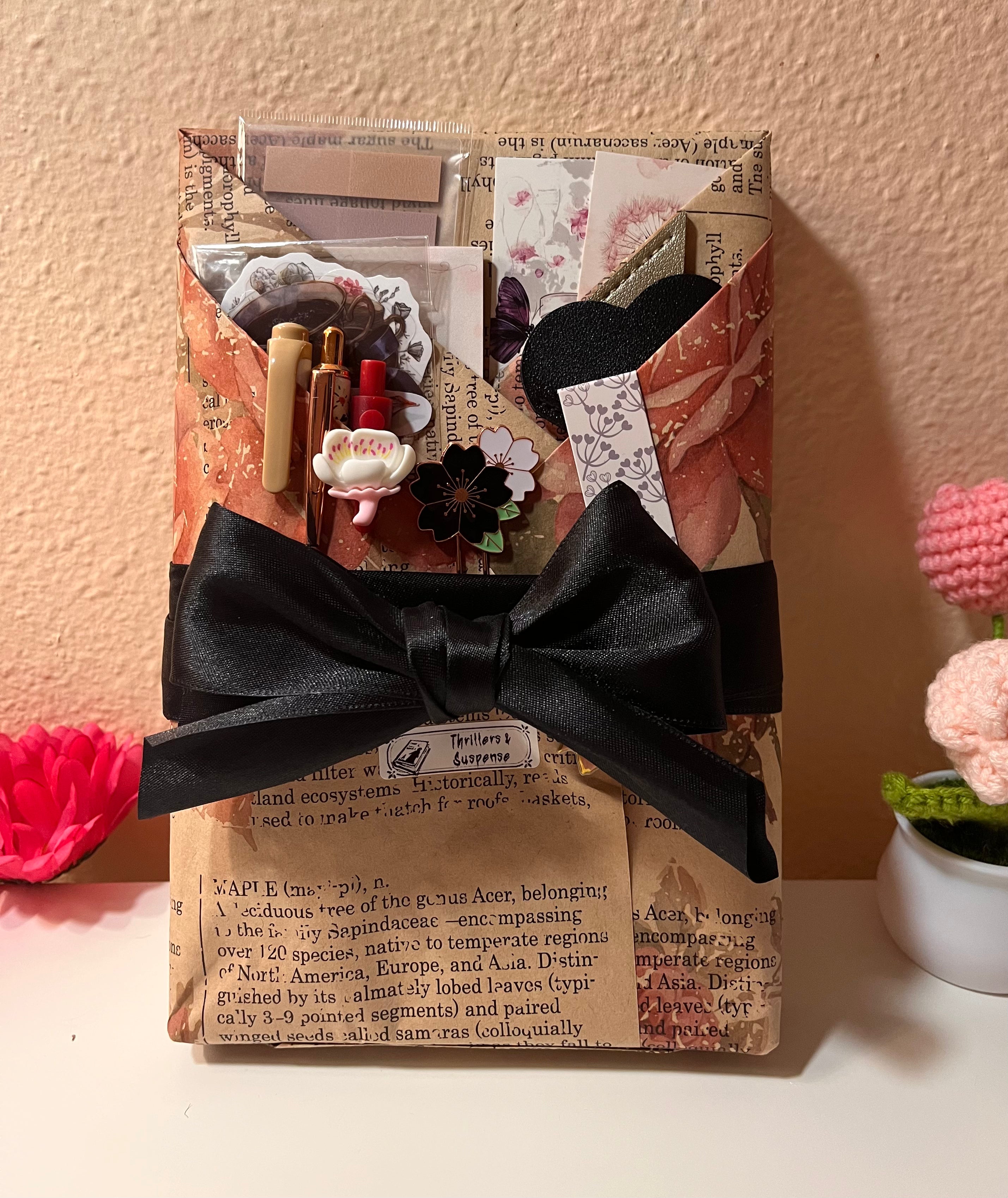 A wrapped Blind date with a book featuring vintage-style bookish stickers, floral patterned paper, decorative book marks, a small label titled “Thrillers and Suspense,” and a large black ribbon bow, set against a warm beige wall with a small pink flower arrangement in the corner