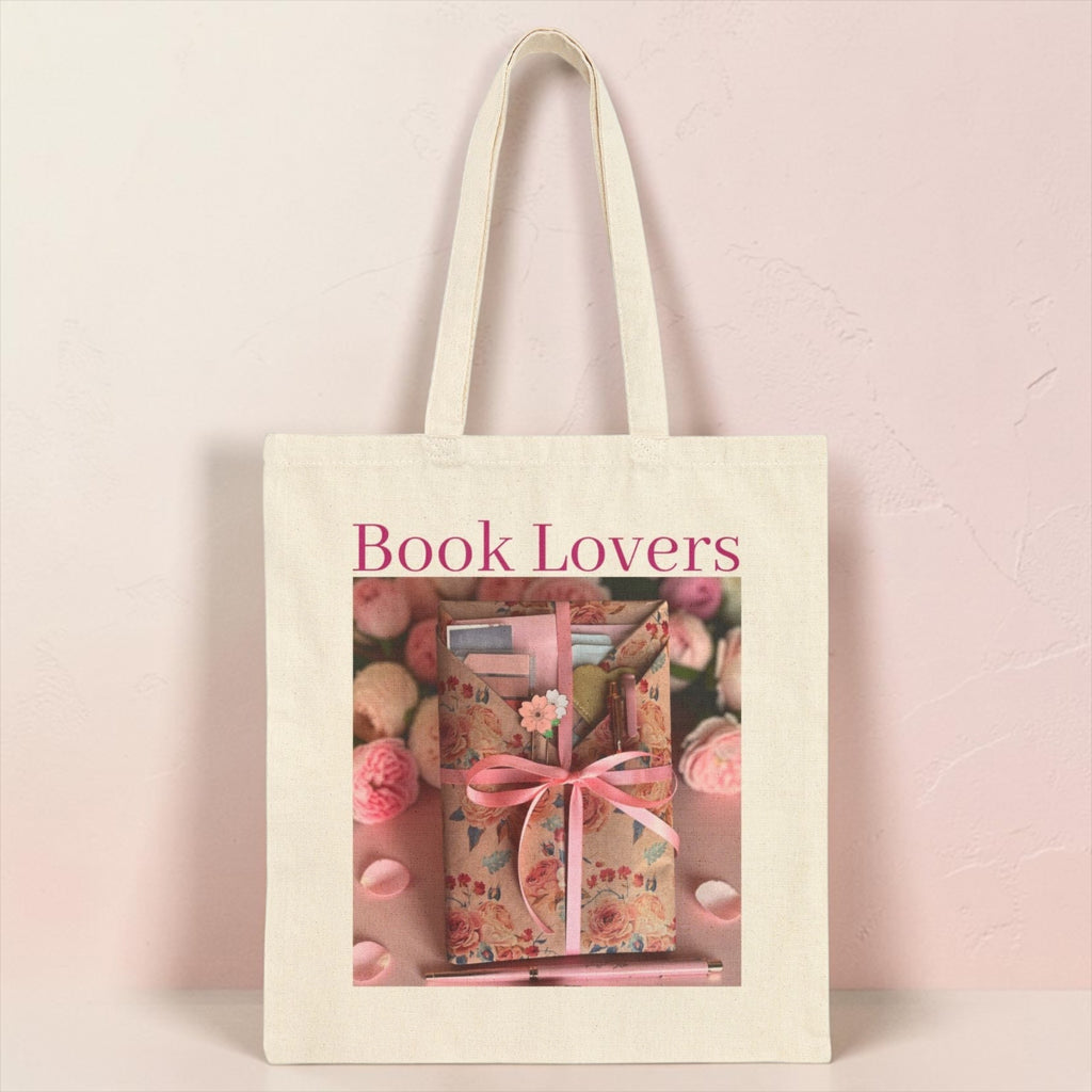 An off-white canvas tote bag with long handles, featuring a printed graphic of a wrapped gift box with a pink ribbon and the words 'Book Lovers' in pink text, set against a pale pink textured background.