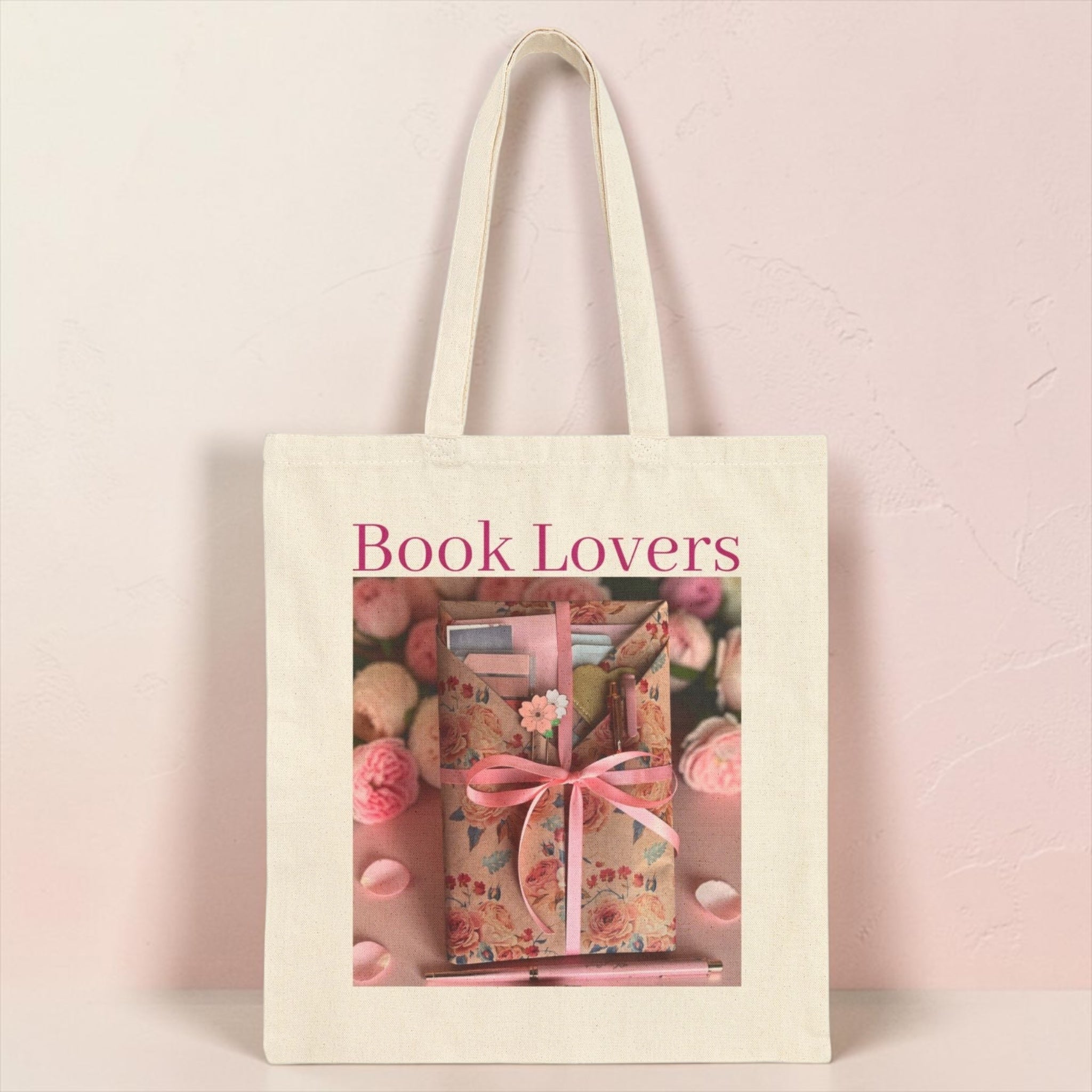 An off-white canvas tote bag with long handles, featuring a printed graphic of a wrapped gift box with a pink ribbon and the words 'Book Lovers' in pink text, set against a pale pink textured background.