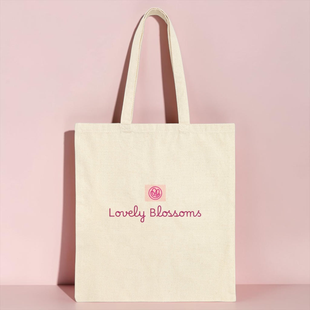 An off-white canvas tote bag with pink embroidered text reading 'Lovely Blossoms' and a small pink circular logo, standing against a solid pink background.
