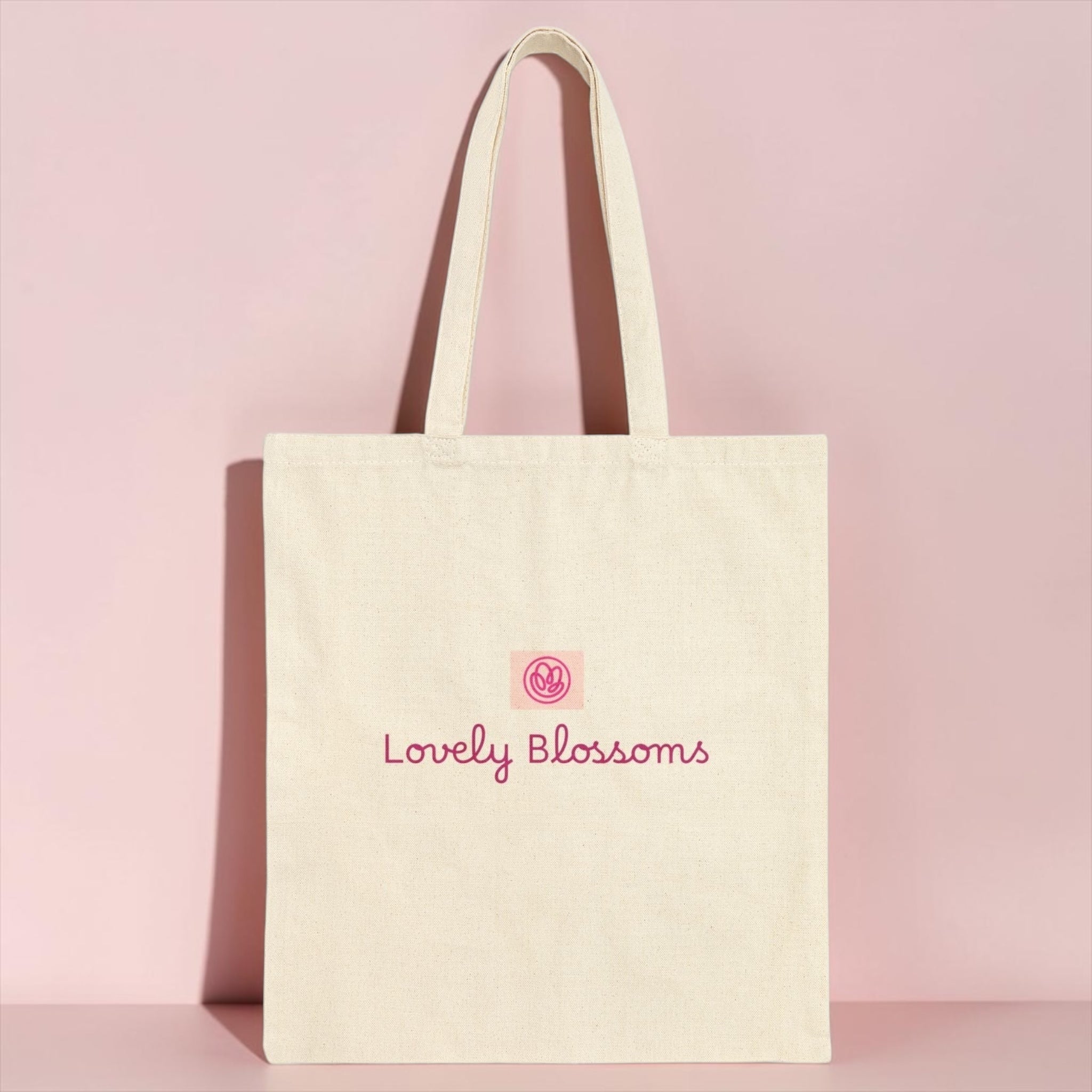 An off-white canvas tote bag with pink embroidered text reading 'Lovely Blossoms' and a small pink circular logo, standing against a solid pink background.