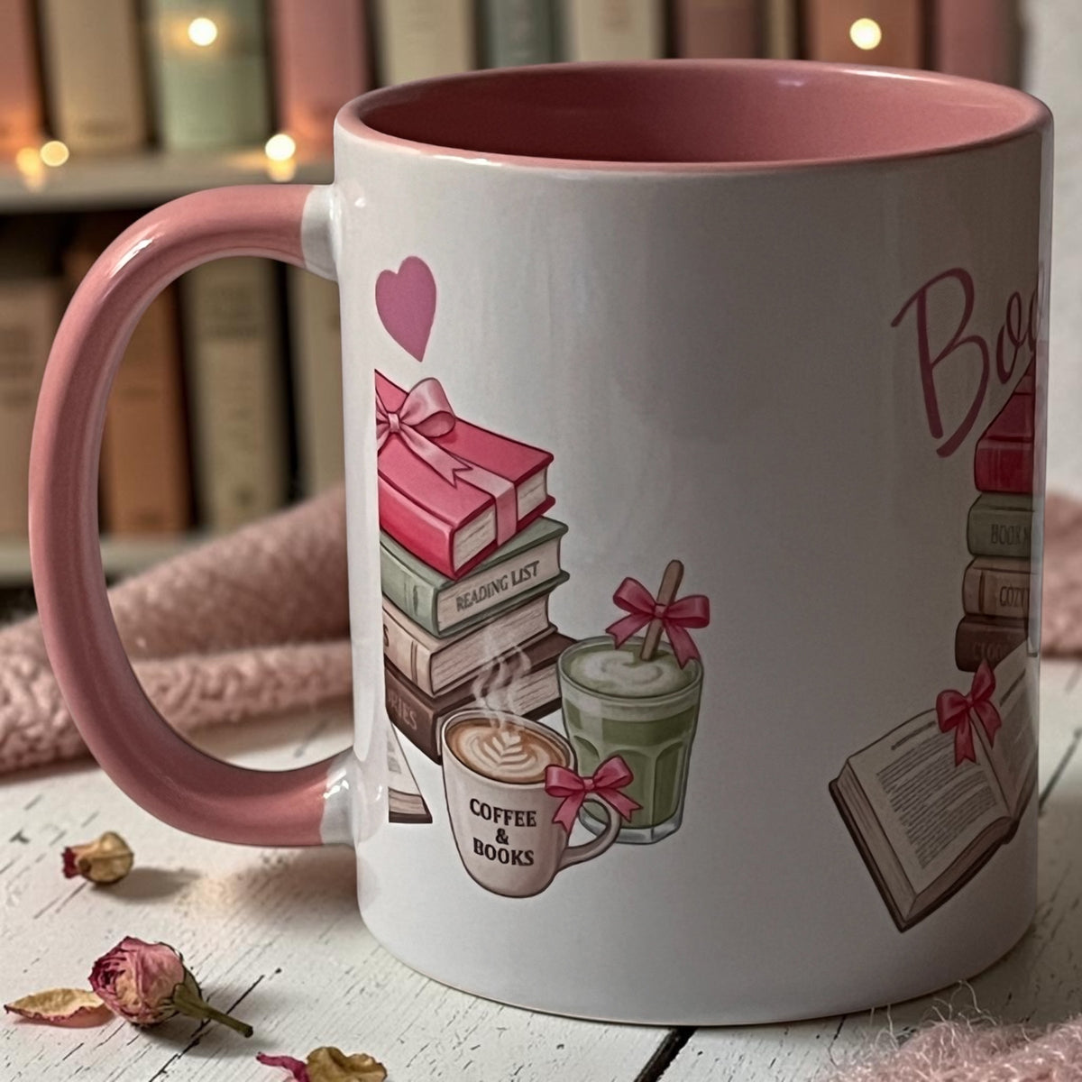 A white ceramic mug with a pink handle and interior, decorated with illustrations of books, a coffee cup, a drink with a straw, and the words 'Books & Coffee'. The mug is photographed on a white wooden surface with dried flowers and a soft pink fabric in the foreground, and blurred bookshelves with warm lights in the background.