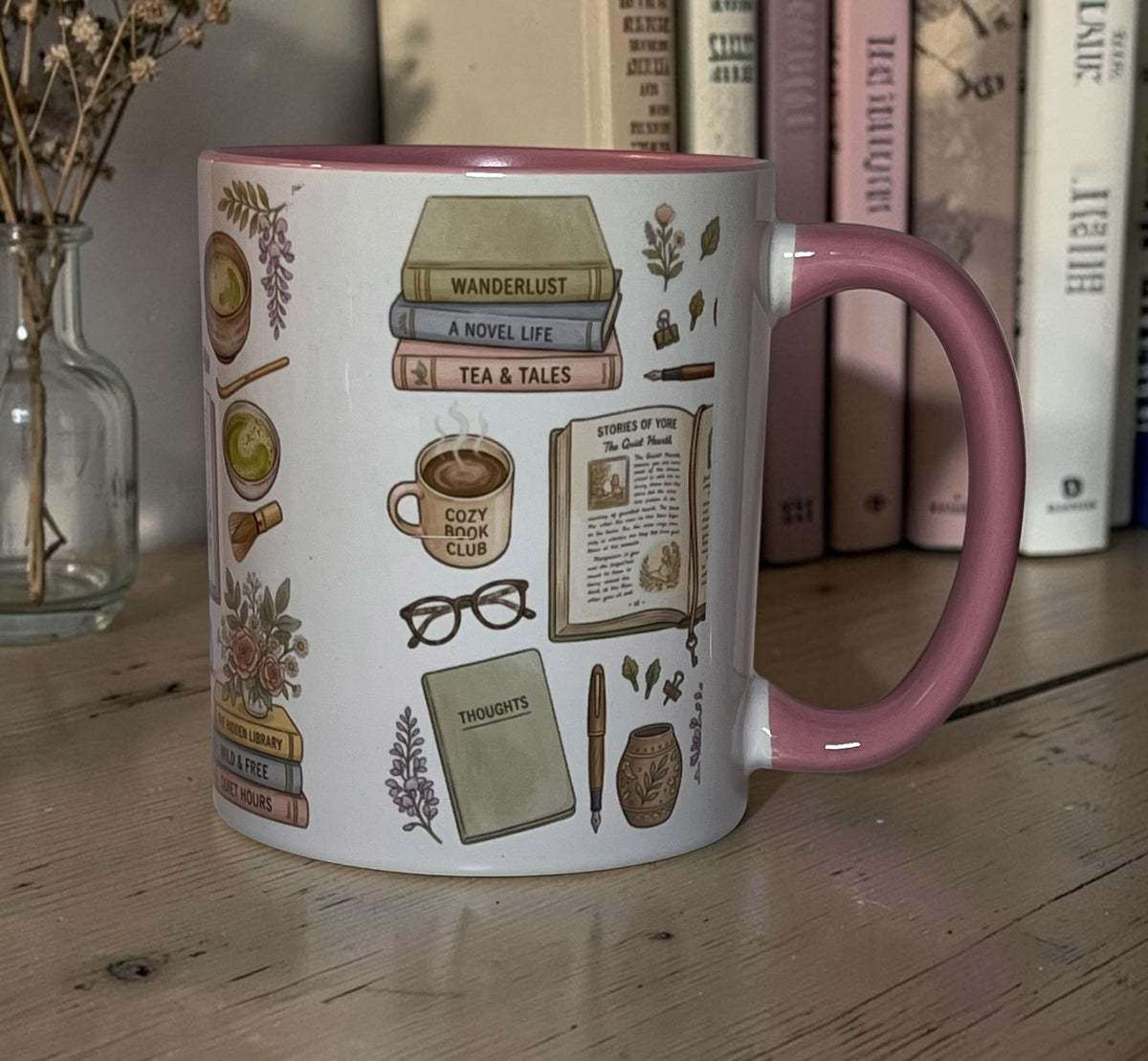 A side view of a 11 ounce ceramic white and pink mug on top of a barn style kitchen table, with a small book shelf in the background. The ceramic mug contains various bookish themed items in the colors of pink, green, brown and purple. Floral patterns are also found on the mug between the bookish themes. 