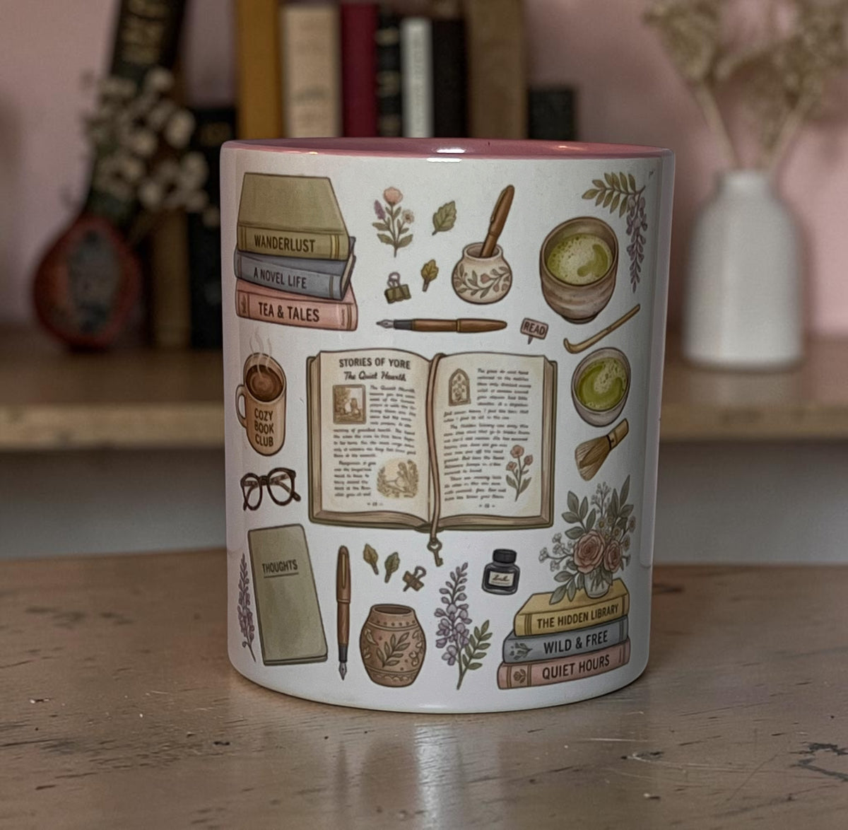 A white ceramic mug with a pink interior rim, decorated with vintage-style illustrations of books, an open book, a pen, glasses, a mug, and botanical motifs. The mug is placed on a wooden table with a blurred background of books and a white vase.