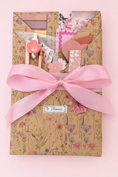 A book hand wrapped in beige floral patterned paper, tied with a large pink satin ribbon, and decorated with small pink and white floral embellishments, a floral pen, bookmarks, floral page paper clip, bookish stickers, pens and a small label reading “Romance” centered at the mid half way of the book. 