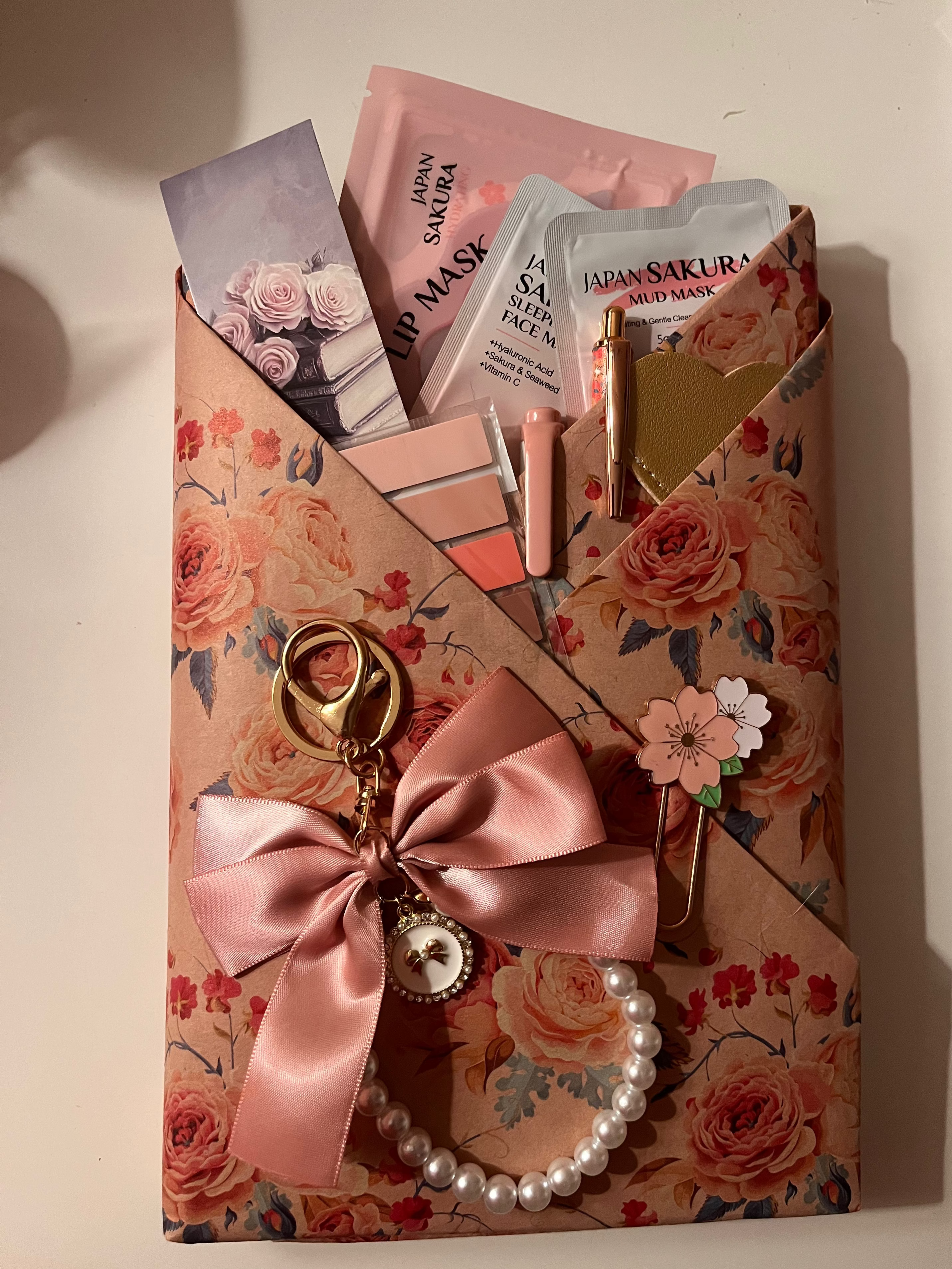 A floral-patterned envelope-style pouch filled with skincare and beauty items, including pink and white mask packets, a gold heart-shaped accessory, and a pink satin bow with pearl beads and a keychain, set against a warm beige background.