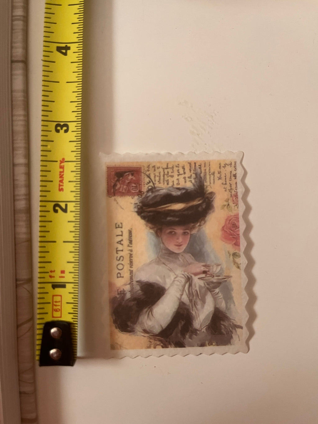 A vintage-style postage stamp featuring an illustration of a woman in a hat, shown next to a yellow Stanley tape measure on a light beige surface.