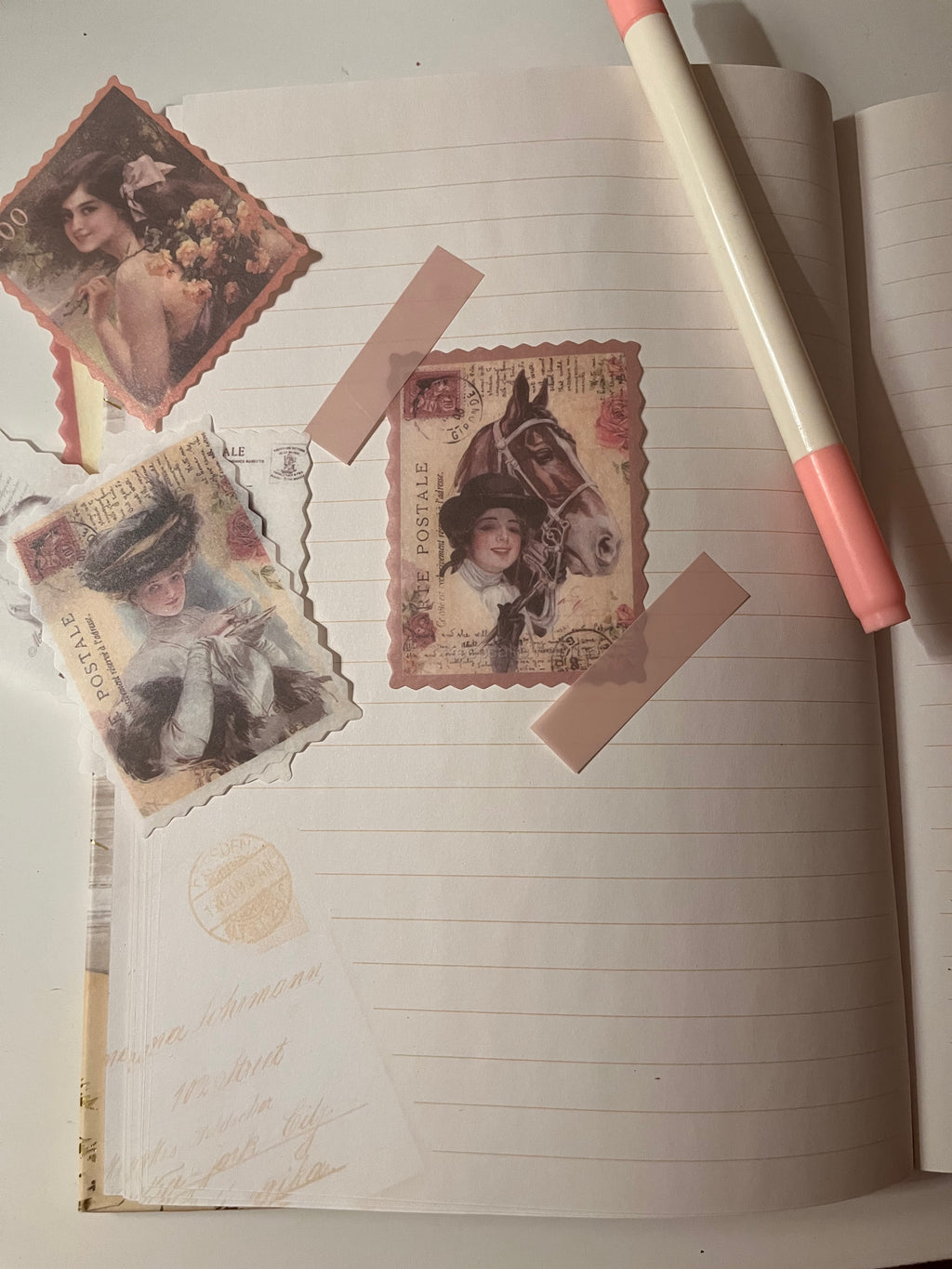 A styled flat-lay of vintage-style decorative paper cutouts and ephemera arranged on an open lined notebook, with a pink-capped pen resting on the right side.